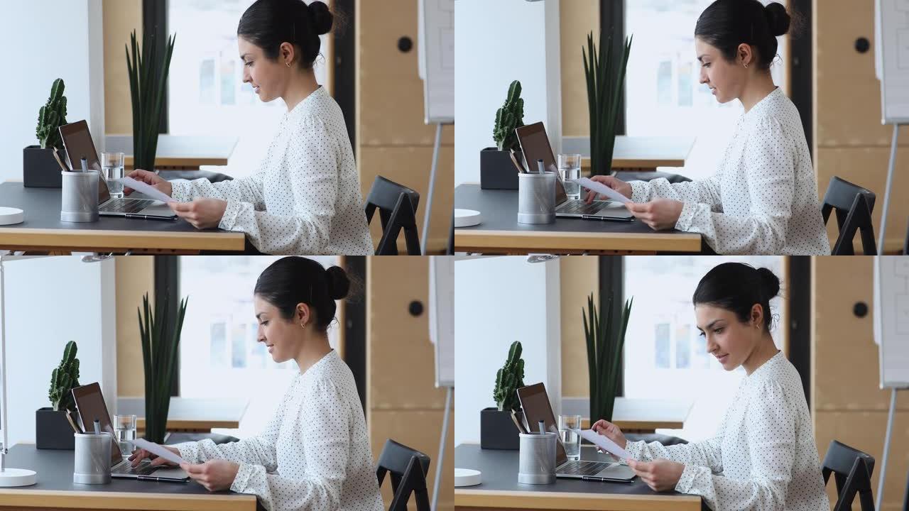 专注的印度年轻女子拿着纸质文件，在电脑上工作。