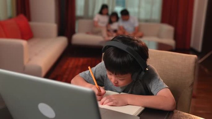 亚洲男孩在网上学习时通过笔记本电脑和耳机做笔记，家人坐在后台的沙发上。电晕病毒爆发，社交距离，儿童教