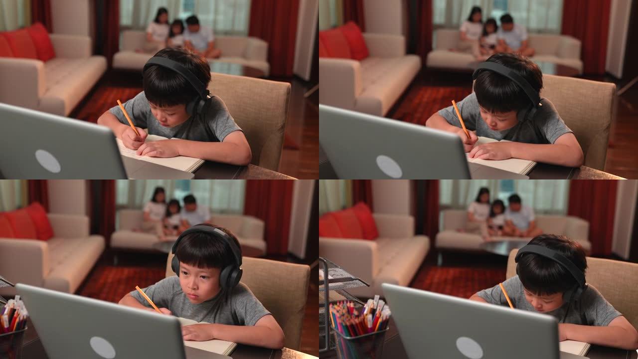 亚洲男孩在网上学习时通过笔记本电脑和耳机做笔记，家人坐在后台的沙发上。电晕病毒爆发，社交距离，儿童教