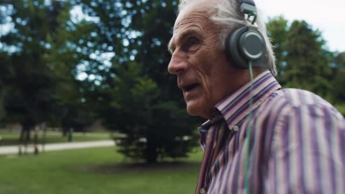 一个活跃的老人在公园里活泼跳舞的肖像，用耳机听音乐。快乐开朗的老年男性，穿着一件彩色衬衫享受生活，伴