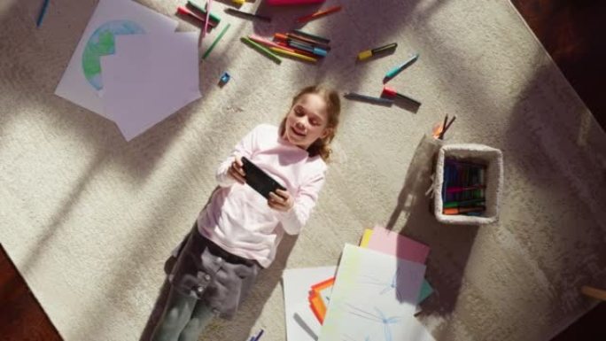 顶视图主页: 快乐的小女孩躺在地板上使用智能手机。快乐的晴天和聪明的微笑孩子玩智能视频游戏，使用手机