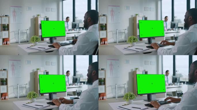 非裔美国医生正在健康诊所使用带有绿屏模拟显示的计算机。穿着白大褂的助理正在医院办公室的桌子后面阅读病