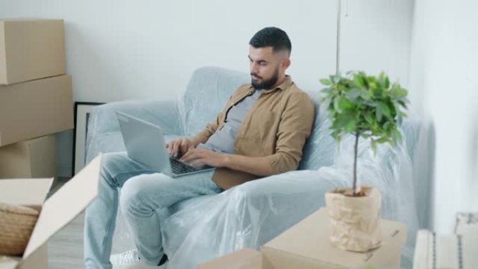 阿拉伯人自由职业者在装满纸板箱的公寓里坐在沙发上的笔记本电脑上工作
