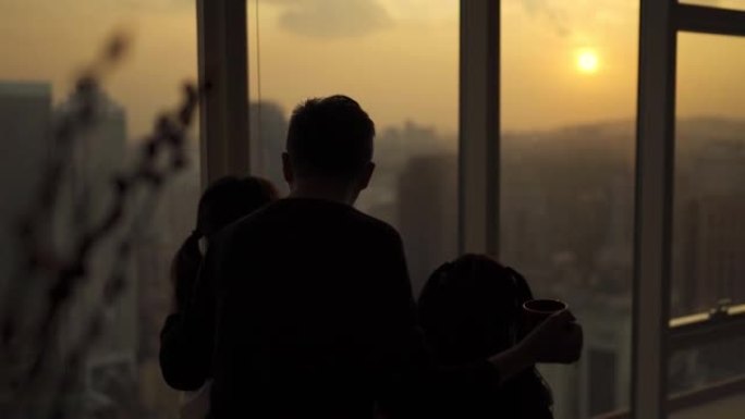 剪影后视亚洲中国家庭，有2个孩子，透过窗户，在吉隆坡市的客厅享受美丽的日落