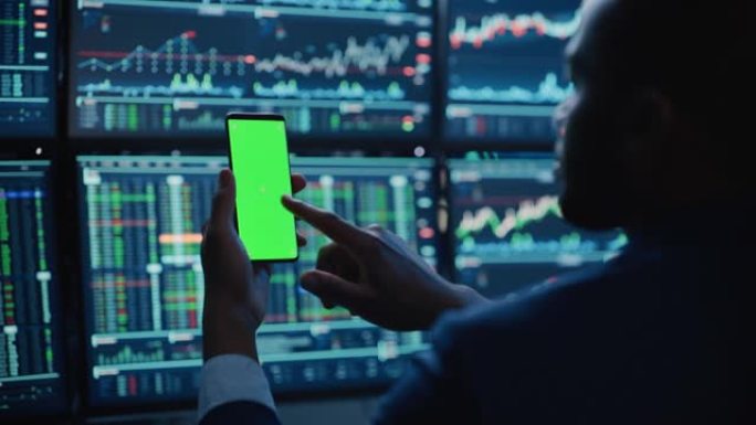 财务分析师使用带有绿屏色度键模拟模板的智能手机，并在带有股票图表的多显示器工作站上工作。商人晚上在投