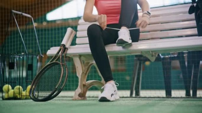 年轻女子坐在长凳上，在室内网球场系鞋带