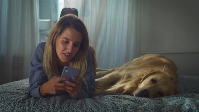 一名女子用智能手机躺在床上，与金毛猎犬狗合影。女狗主人与她的熟睡宠物共度时光，同时保持联系，聊天。检