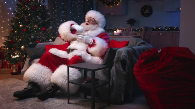 圣诞老人坐在装饰精美的客厅里过圣诞节，坐在灰色的沙发上看电视，吃着爆米花。圣诞老人热情地将爆米花倒入