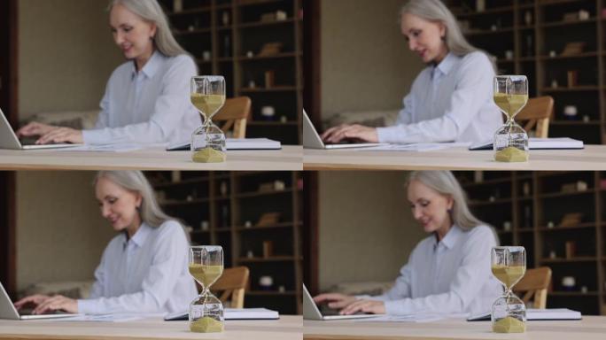 成熟的女人在笔记本电脑键盘上打字用沙镜计时