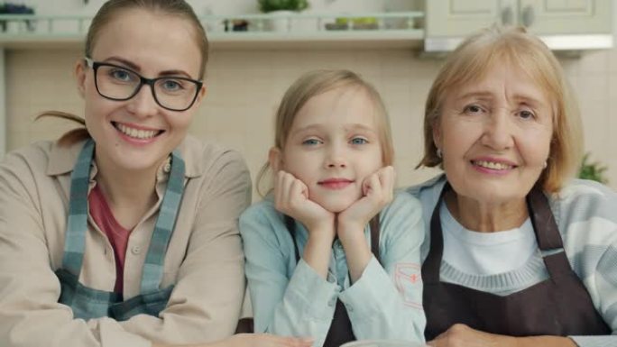 奶奶、妈妈和孩子微笑的肖像坐在厨房里穿着围裙看着相机