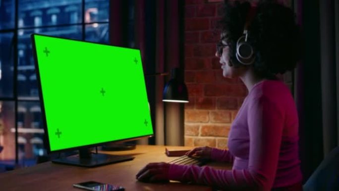 戴着耳机的年轻漂亮女性在台式电脑上在家工作，绿屏模拟显示。有创造力的女人检查社交媒体，浏览互联网。从