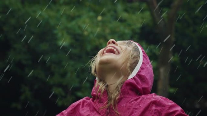 一个穿着粉红色雨衣的快乐小女孩在雨中快乐地站着，被大自然和树木包围着。女孩微笑着，在雨天独自在外面玩