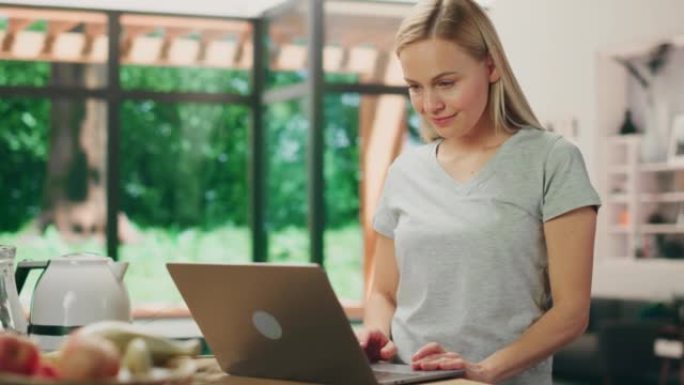 美丽的年轻成年女性肖像，金发碧眼，穿着灰色v领t恤，站在客厅时使用笔记本电脑。在光明之家在家工作的成