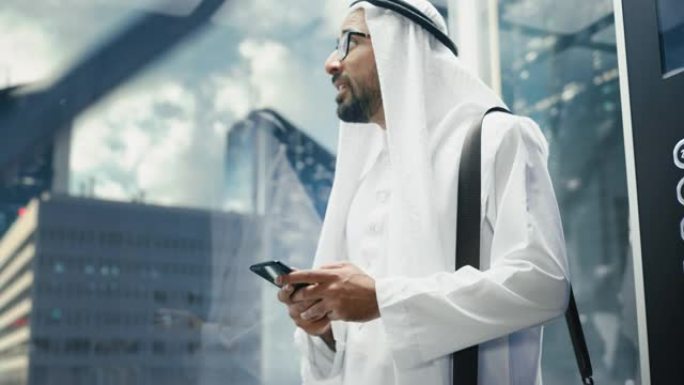 成功的穆斯林商人在传统的白色坎杜拉骑着玻璃电梯到现代商务中心的办公室。男人使用智能手机。沙特、阿联酋