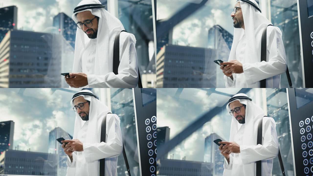 成功的穆斯林商人在传统的白色坎杜拉骑着玻璃电梯到现代商务中心的办公室。男人使用智能手机。沙特、阿联酋