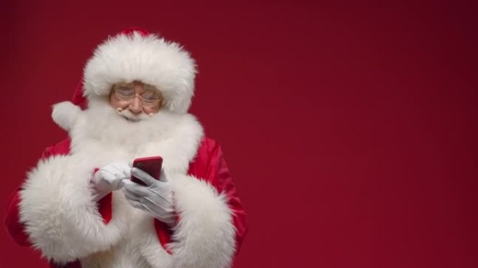 一个开朗的圣诞老人正在热情地在网上购物，在红色智能手机上选择一些东西，站在红色背景下，有一个链接的地