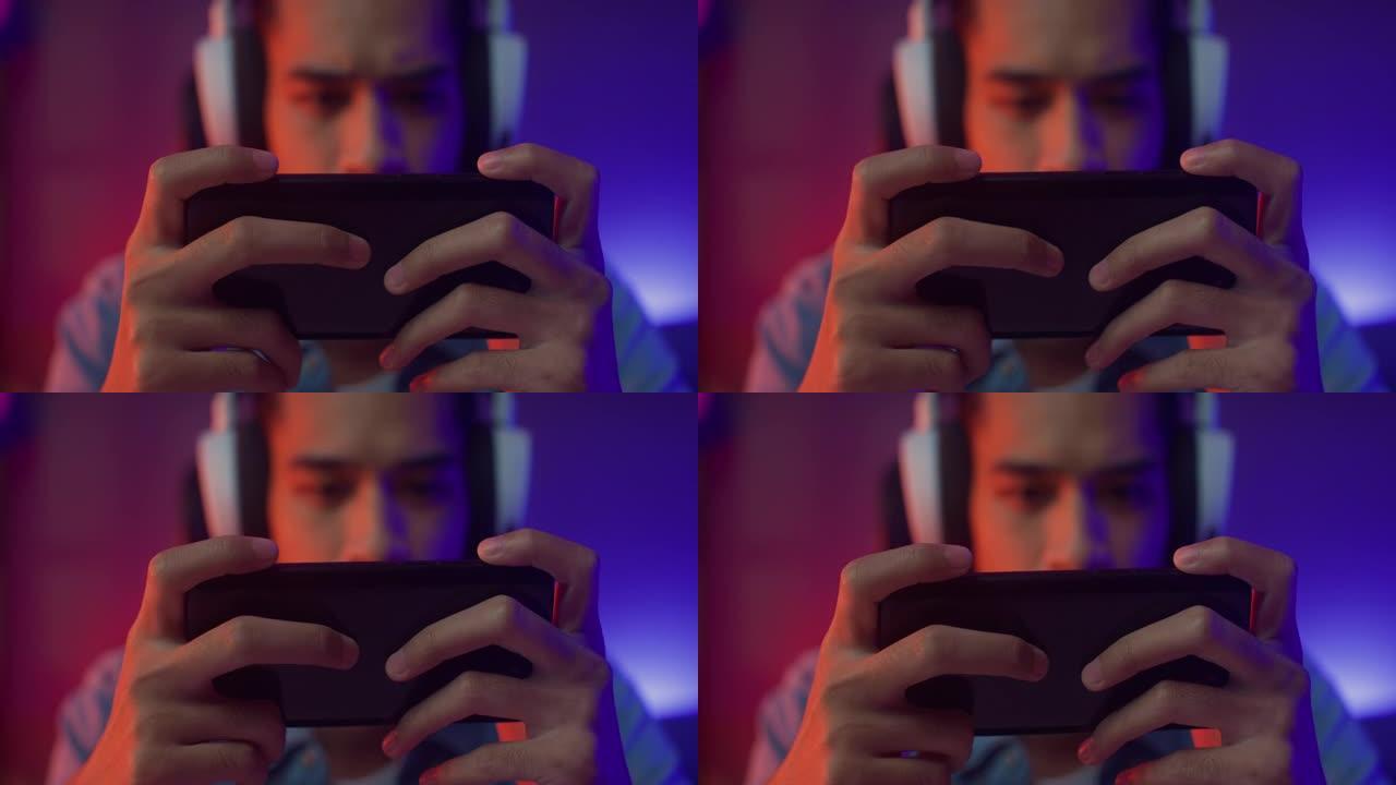快乐亚洲男子游戏玩家戴着耳机比赛在线玩视频游戏，晚上在客厅用智能手机彩色霓虹灯现代房子。
