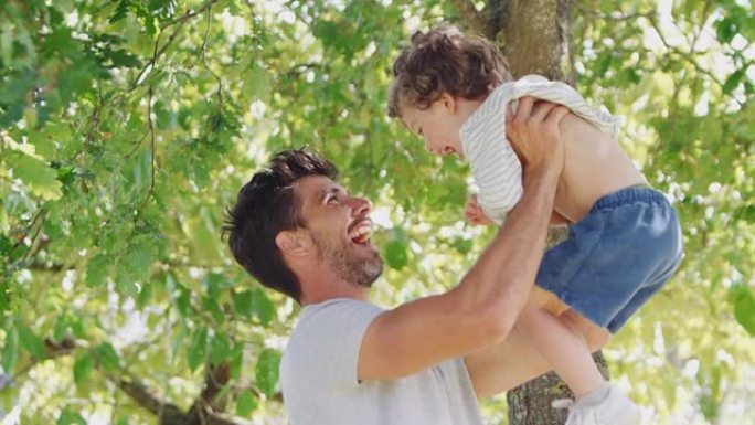 慈爱的父亲和儿子在公园里玩得开心，在绿树成荫的背景下将他抱在空中