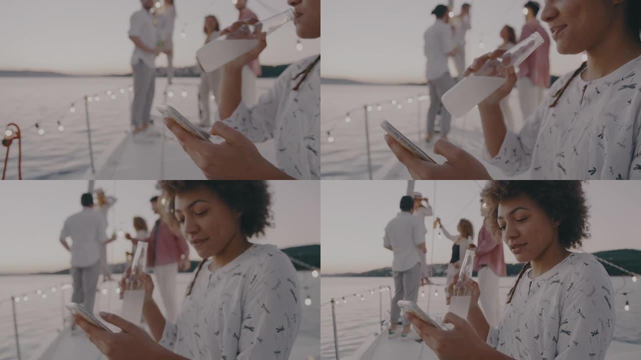 SLO MO Young woman喝酒并使用她的智能手机，而她的朋友聚集在船的背景中