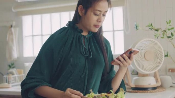 美丽的女人在厨房吃沙拉时使用智能手机。
