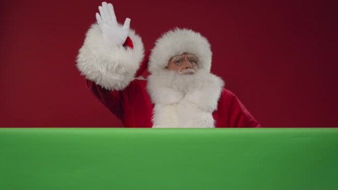 欢快的圣诞老人在红色背景上的绿色屏幕后面的框架中前进，向相机挥手并离开