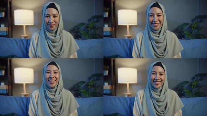 迷人开朗的年轻亚洲穆斯林美女戴着头巾，晚上在家里舒适的客厅微笑着看着相机。女孩伊斯兰检疫生活方式概念