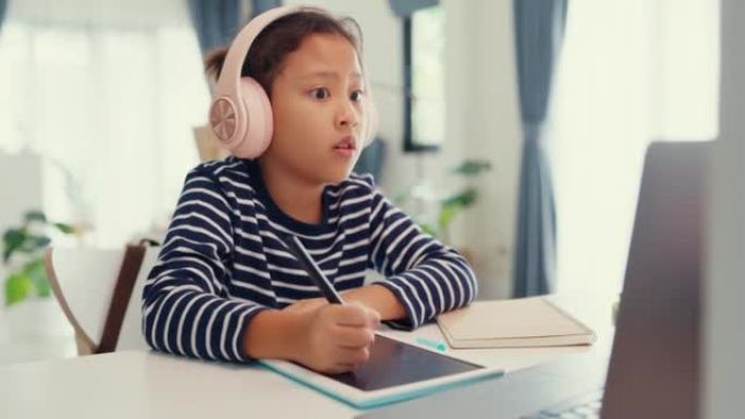 亚洲蹒跚学步的女孩戴着耳机坐在带记事本的书桌前，使用魔术笔专注于写作业，在家从笔记本电脑上计算数字数