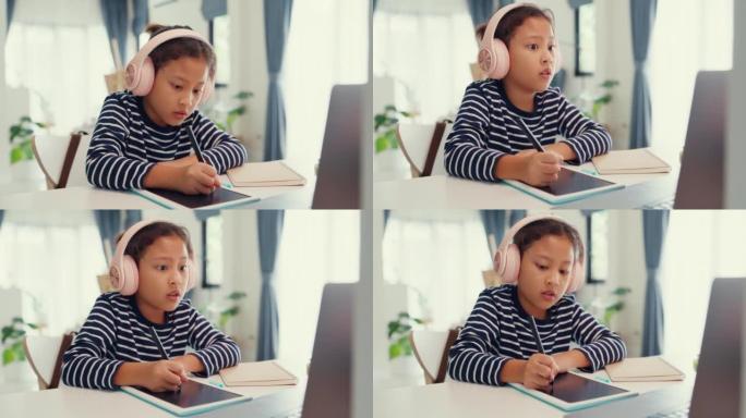 亚洲蹒跚学步的女孩戴着耳机坐在带记事本的书桌前，使用魔术笔专注于写作业，在家从笔记本电脑上计算数字数
