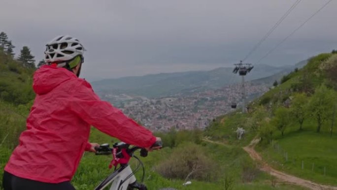 穿着红色夹克和头盔的女人从骑自行车旅行中休息一下，站在山顶上，可以看到萨拉热窝，背景是缆车