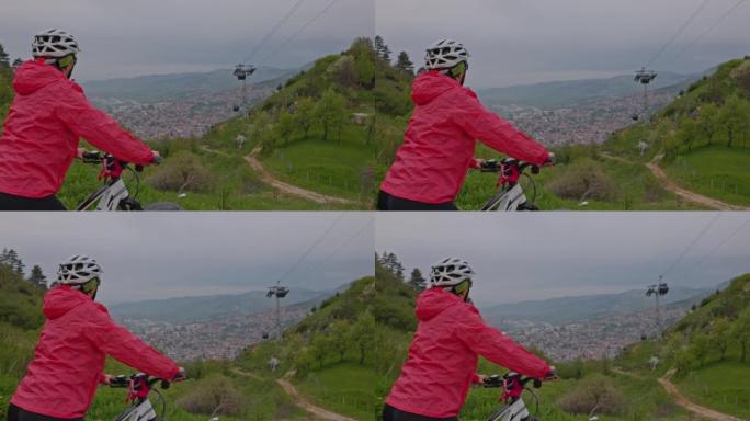 穿着红色夹克和头盔的女人从骑自行车旅行中休息一下，站在山顶上，可以看到萨拉热窝，背景是缆车