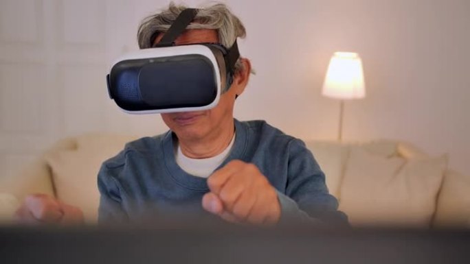 幸福的亚洲老年男性62岁戴着虚拟现实耳机和手势，而玩在线游戏使用电脑电脑坐在他的书桌上在家。高级技术