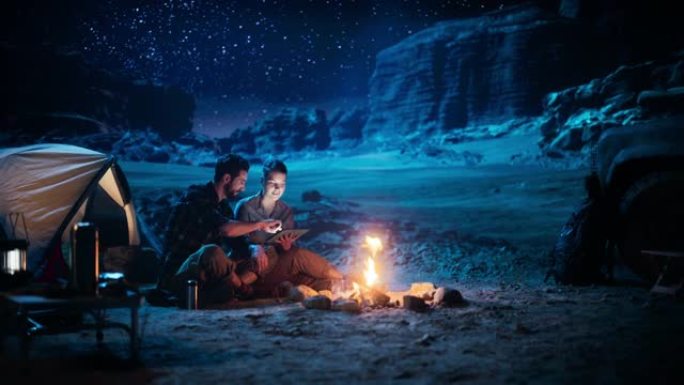 快乐情侣大自然晚上在峡谷露营，使用数字平板电脑，坐在篝火旁。两个旅行的人玩得开心，微笑，在社交媒体上
