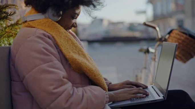 年轻的黑人妇女在城市的长凳上使用笔记本电脑时微笑
