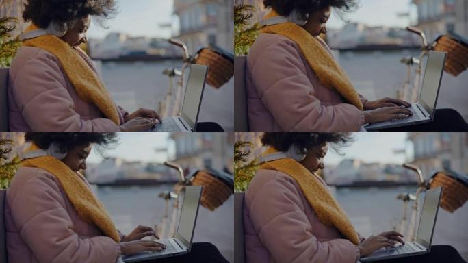 年轻的黑人妇女在城市的长凳上使用笔记本电脑时微笑