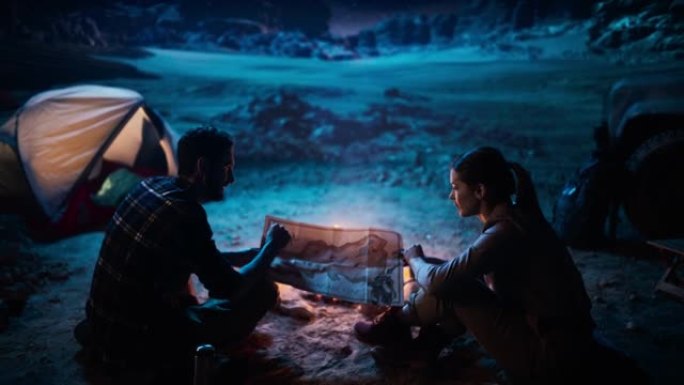 幸福的夫妇晚上在峡谷露营，坐在篝火旁，用地图规划徒步旅行路线。两个旅行徒步旅行者交谈，探索自然，环游