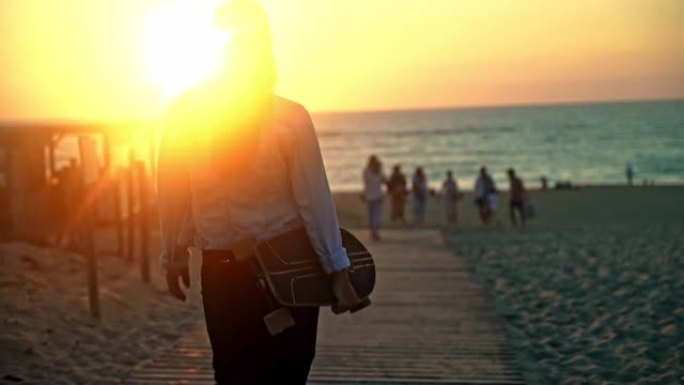 日落时，一头棕色头发的年轻女子在手臂下背着滑板走向海滩