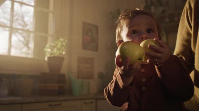 电影拍摄的快乐微笑可爱的好奇的蹒跚学步的小男孩吃新鲜的充满维生素的生物苹果，而他的母亲在家里的厨房里