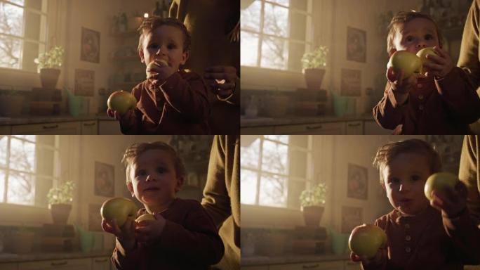 电影拍摄的快乐微笑可爱的好奇的蹒跚学步的小男孩吃新鲜的充满维生素的生物苹果，而他的母亲在家里的厨房里