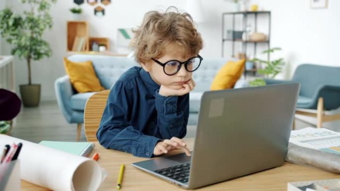 聪明的小男孩戴着眼镜在公寓的桌子上使用笔记本电脑