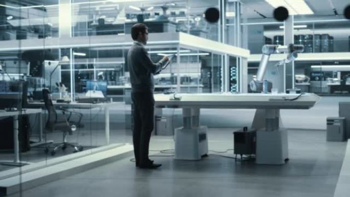 工业机器人工程师来到一家高科技工厂的带机械臂的桌子前。男性科学家使用平板电脑来操纵和编程机器人来拾取