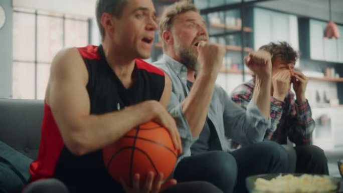 在家里，三个快乐的篮球迷坐在电视上的沙发上观看比赛，庆祝得分，运动队冠军的胜利。一群朋友为最喜欢的俱