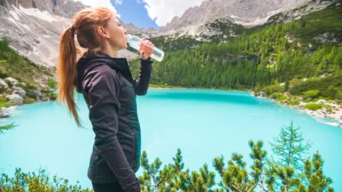 年轻女子站在雄伟的绿松石山湖前，用可重复使用的水瓶补水