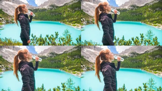 年轻女子站在雄伟的绿松石山湖前，用可重复使用的水瓶补水