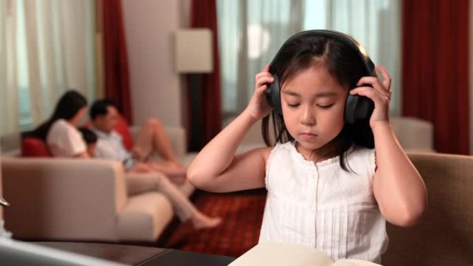 亚洲女孩在网上学习时通过笔记本电脑和耳机做笔记，家人坐在沙发上的背景。社交距离，儿童教育，自学，电子