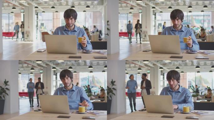 商人在现代办公室里用热饮在办公桌上的笔记本电脑上工作，背景是同事