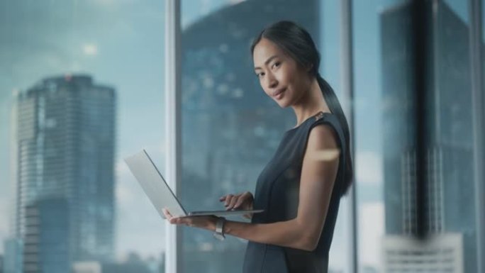 一位穿着时髦的黑色连衣裙的亚洲女商人的美丽肖像，使用笔记本电脑，在城市办公室的窗户旁边摆姿势。自信的