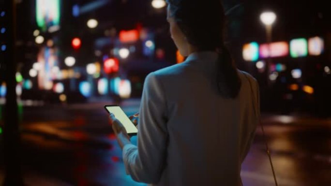 美丽的女人在智能手机上在线购物，同时穿过充满霓虹灯的夜晚城市街道。女性使用手机、电子商店、互联网服装