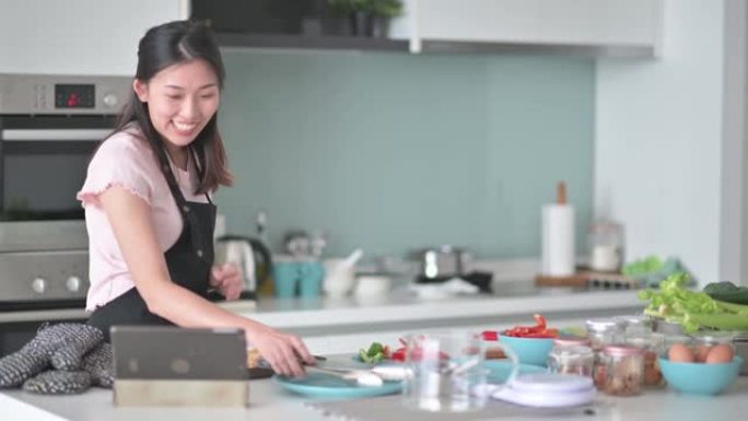 周末在厨房柜台，戴着防护手套的亚洲中国女性拿着一盘蛋挞看着家里微笑的数字平板电脑