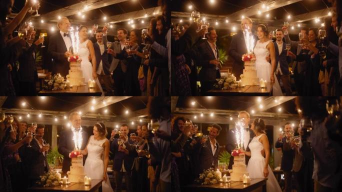美丽的新娘和新郎在晚宴上与多民族朋友庆祝婚礼。新婚夫妇提议为幸福的婚姻干杯，站在餐桌上，带着蛋糕和烟