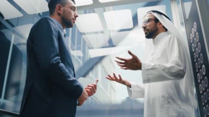 商人在现代商务中心乘坐玻璃电梯到办公室时与阿拉伯投资伙伴交谈。国际企业合伙人握手并达成协议。